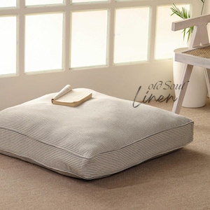 Puede incluir: Un cojín de suelo grande y cuadrado con tela de rayas blancas y negras. El cojín está hecho de lino y está sentado en una alfombra marrón claro. Un libro está descansando sobre el cojín.