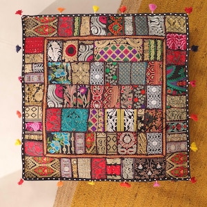 May include: A square black cushion with a patchwork design. The cushion is made of various fabrics in shades of red, blue, green, gold, and purple. The cushion has a fringe border with tassels.