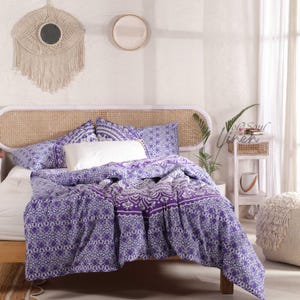 May include: Purple and white mandala patterned duvet cover and pillowcases on a bed with a white wicker headboard. The bedding is on a white bed frame with a white bedside table and a white stool.