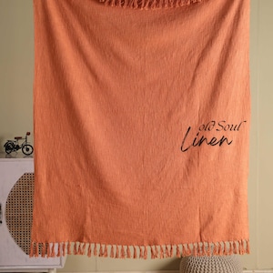 Op de afbeelding: Een koraalkleurige linnen sprei met franjes langs de boven- en onderranden. De woorden "old Soul Linen" staan in cursieve letters rechtsonder op de deken. De deken hangt.