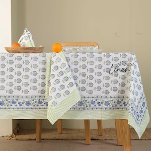 Puede incluir: Mantel blanco con un estampado floral azul y un borde a rayas verdes. Una servilleta a juego y un cuenco de madera con naranjas están sobre la mesa. El mantel está sobre una mesa de madera.