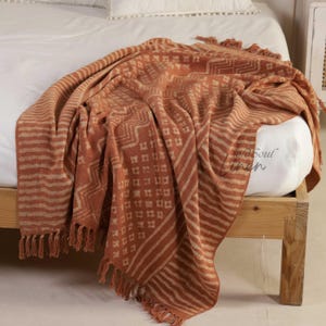 Könnte beinhalten: Eine terracottafarbene Decke mit geometrischem Muster und Fransen. Die Decke hat ein sich wiederholendes Design aus Quadraten, Zickzacklinien und Streifen in einer helleren Beigefarbe. Die Decke ist über ein Bett drapiert.