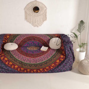 Pode incluir: Uma tapeçaria mandala de cor roxo escuro e vermelho com padrões florais intrincados e um acento dourado. A tapeçaria está drapeada sobre um sofá com duas almofadas brancas.