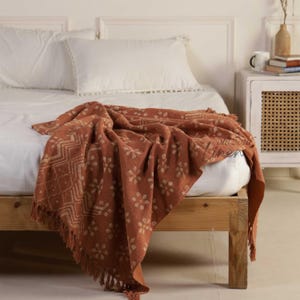 Op de afbeelding: Een terracotta kleurige sprei met een bloemen- en geometrisch patroon over een bed. De sprei heeft een gefranjerde rand en is gemaakt van een zachte, getextureerde stof. Het bed heeft een houten frame en wit beddengoed.