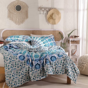 May include: A white bed with a blue and white patterned duvet cover and matching pillowcases. The bed has a white wicker headboard and is styled with a woven wall hanging and a straw hat.