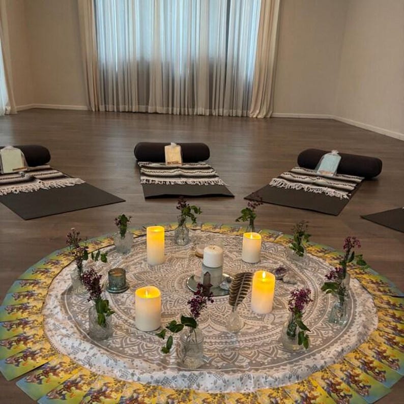 May include: A white circular tablecloth with a mandala pattern, surrounded by lit candles and small vases with flowers. There are tarot cards spread out on the floor around the tablecloth.