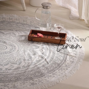 May include: A white round mandala rug with a gray pattern and fringe trim. The rug is on a white floor with a wooden tray, a glass of water, and a glass pitcher.