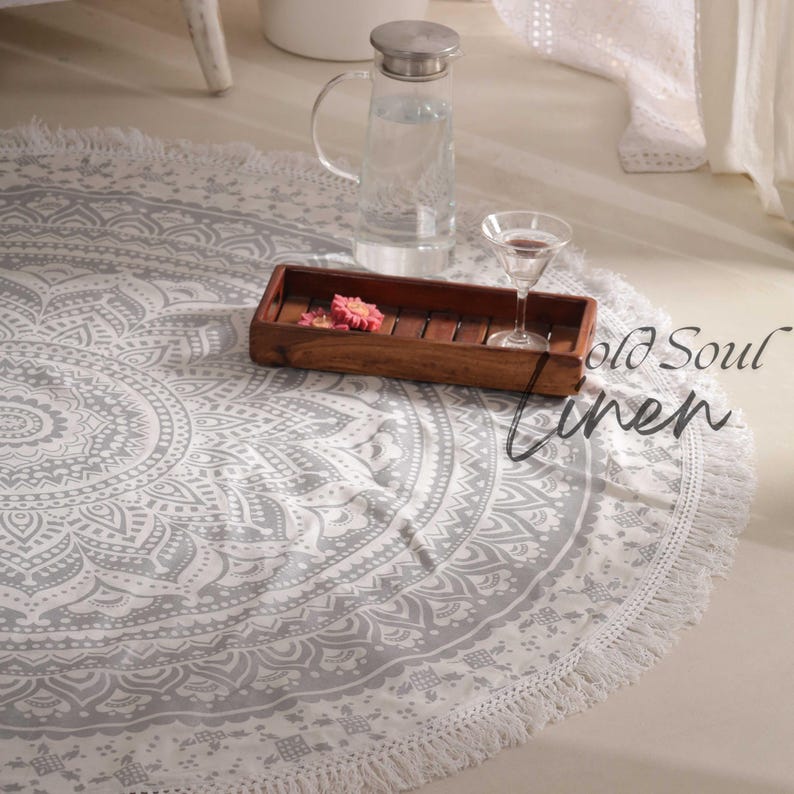 May include: A white round mandala rug with a gray pattern and fringe trim. The rug is on a white floor with a wooden tray, a glass of water, and a glass pitcher.