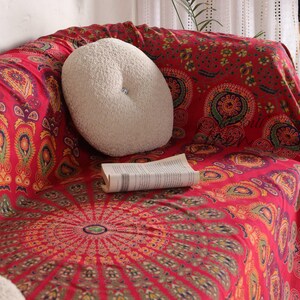Puede incluir: Una manta de tiro con estampado de mandala rojo y dorado con una almohada redonda blanca en un sof&aacute;.
