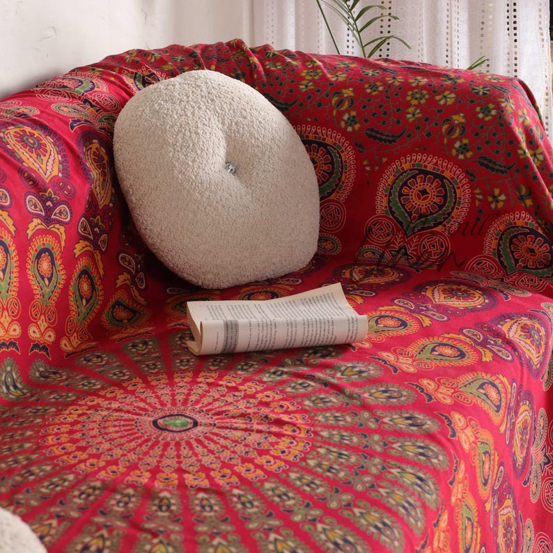 Puede incluir: Una manta de tiro con estampado de mandala rojo y dorado con una almohada redonda blanca en un sof&aacute;.