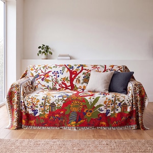 May include: A sofa covered in a white fabric throw featuring a vibrant, multi-colored botanical and animal print with red, yellow, blue, and green hues. The throw displays stylized trees, birds, and small animals. Two solid-colored pillows rest on top.