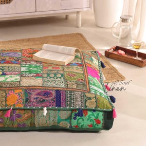Puede incluir: Un gran cojín de suelo cuadrado con un diseño de patchwork. El cojín está hecho de varias telas en tonos de verde, rosa, azul y dorado. El cojín tiene un borde de flecos y borlas.