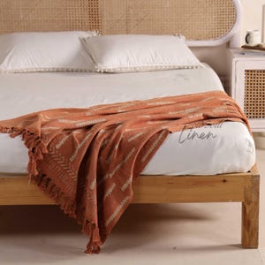 Puede incluir: Una manta de color naranja y crema con un patr&oacute;n geom&eacute;trico y flecos est&aacute; sobre una cama. La cama tiene s&aacute;banas blancas y un marco de madera. El cabecero es de mimbre tejido.