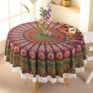 Op de afbeelding: Een ronde, bedrukte tafelkleed met een pauwenveerontwerp in paars, oranje, groen en rood. Het tafelkleed heeft een witte franjebies en staat op een houten tafel. Bloemen en een dienblad met een kopje staan op de tafel.
