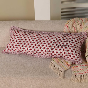 May include: Rectangular decorative pillow with a red and white geometric pattern. The pillow has a ruffled edge and a script design in the center. It rests on a beige sofa with a fringed throw blanket.