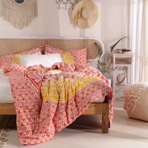 Op de afbeelding: Een bed in boho-stijl met een wit hoofdeinde en een kleurrijk, gepatroneerd dekbedovertrek. Het dekbedovertrek heeft een mandala-ontwerp in tinten roze, oranje en geel. Het bed is gestyled met witte kussens en een geweven hoofdeinde.
