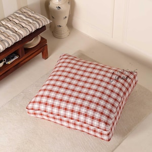 May include: A square floor cushion with a red and white plaid pattern. The cushion is placed on a light-colored rug. A wooden bench with a striped cushion and a decorative vase are in the background.