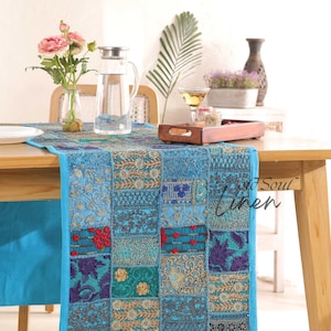 Op de afbeelding: Een blauwe patchwork tafelrunner met gouden borduurwerk. De runner is gemaakt van verschillende stoffen en heeft een verscheidenheid aan patronen. Hij ligt op een houten tafel met een glazen kan, een glas wijn en een houten dienblad.