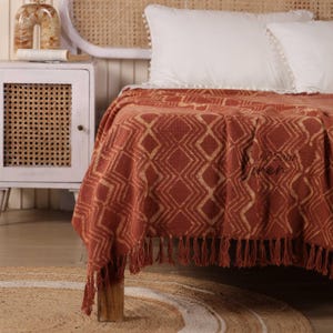 Könnte beinhalten: Terracottafarbene Decke mit geometrischem Rautenmuster und Fransenbesatz. Die Decke ist über einem Bett mit weißen Kissen und einem gewebten Kopfteil drapiert. Der Text "Soul Linen" ist sichtbar.