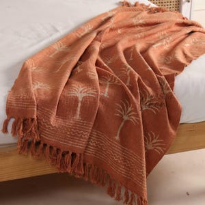 Puede incluir: Una manta naranja con un estampado de palmeras y flecos. La manta está drapeada sobre una cama, mostrando el diseño detallado y la textura. El diseño de palmera es de un tono más claro, creando un contraste.