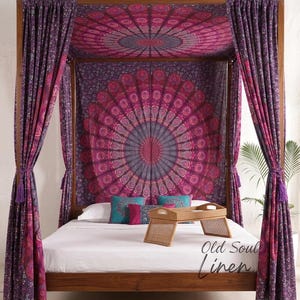 Puede incluir: Una cama con dosel de madera con un cabecero de tapiz de mandala y cortinas a juego en tonos morados y rosas. La cama está hecha con ropa de cama blanca y almohadas decorativas. Una bandeja tejida está sobre la cama y el texto "Old Soul Linen" es visible.