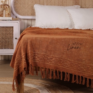 Könnte beinhalten: Eine rostfarbene gewebte Tagesdecke mit geometrischem Muster und Fransenbesatz. Die Decke ist über einem Bett mit weißen Kissen drapiert. Der Text "Soul Linen" ist sichtbar.