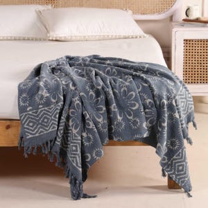 May include: A blue throw blanket with a floral and geometric pattern draped over a wooden bed frame. The blanket has a fringe trim and is made of a soft, textured fabric. White pillows with pom-pom trim are on the bed.