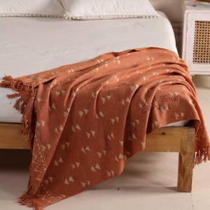 Op de afbeelding: Terracotta-kleurige sprei met een geometrisch driehoekpatroon en franjes. De sprei is over een bed gedrapeerd en toont de textuur en het ontwerp. De sprei is gemaakt van een zacht materiaal.
