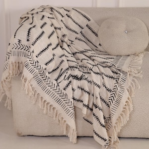 Puede incluir: Una manta crema con un patr&oacute;n geom&eacute;trico negro y flecos est&aacute; sobre un sof&aacute; beige. La manta presenta un dise&ntilde;o de zigzag repetido y un borde con l&iacute;neas verticales y formas de flecha. Una almohada redonda con un bot&oacute;n tambi&eacute;n est&aacute; en el sof&aacute;.