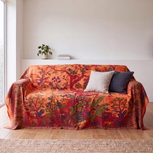 Może przedstawiać: Sofa przykryta jaskrawopomarańczowym kocem ze szczegółowym, wielokolorowym nadrukiem roślinnym i zwierzęcym. Koc przedstawia stylizowane drzewa, ptaki i małe stworzenia w odcieniach czerwieni, błękitu i zieleni. Na wierzchu leżą dwie poduszki dekoracyjne.