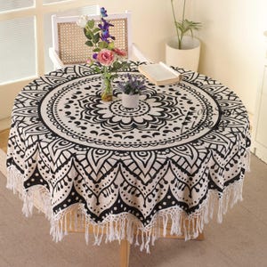 May include: A round white tablecloth with a black mandala design and fringe trim. The tablecloth is draped over a round table with a wooden base. There are flowers and a book on the table.