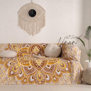 Könnte beinhalten: Ein Sofa, das mit einem gelben, braunen und weißen Mandala-Musterstoff bedeckt ist. Über dem Sofa hängt ein Makramee-Wandbehang mit einem kreisförmigen Spiegel. Die Worte "old Soul linen" stehen auf der rechten Seite des Sofas.