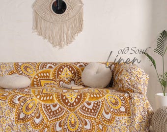 Puede incluir: Un sofá cubierto con una tela con estampado de mandala amarillo, marrón y blanco. Una decoración de pared de macramé con un espejo circular cuelga sobre el sofá. Las palabras "old Soul linen" están escritas en el lado derecho del sofá.