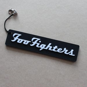 Puede incluir: Una etiqueta rectangular negra con el texto blanco "Foo Fighters". La etiqueta está unida a una cuerda negra con un pequeño cierre plateado. La etiqueta está sobre una superficie marrón claro.