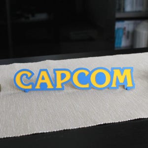 Könnte beinhalten: Eine Präsentation mit einer Link-Figur, einer rosa Kirby-Figur und einem blau-gelben "CAPCOM"-Schild. Die Figuren stehen auf einer neutralfarbenen Oberfläche vor einem dunklen Hintergrund.
