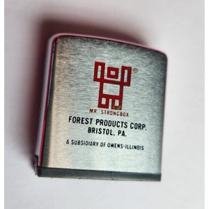 May include: A silver metal tape measure with red accents and a red logo. The text "MR. STRONGBOX" and "FOREST PRODUCTS CORP. BRISTOL, PA." are printed on the front. The tape measure is a subsidiary of Owens-Illinois.