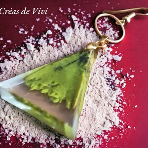 May include: A triangular green and brown resin earring with a gold hook. The earring is on a red background with white powder.