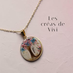 Peut inclure: Un collier à chaîne dorée avec un pendentif rond orné d'un motif d'arbre, de fleurs séchées roses et bleues, de flocons dorés et du prénom Victor en écriture noire. Le texte Les créas de Vivi est visible dans le coin supérieur droit.