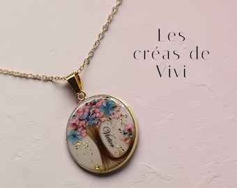 May include: A gold necklace with a circular pendant. The pendant has a tree design with pink and blue flowers, gold flakes, and the name "Victor". The text "Les créas de Vivi" is visible in the background.