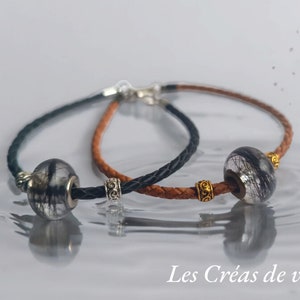 Könnte beinhalten: Zwei geflochtene Lederarmbänder, eines in Schwarz und eines in Braun, mit jeweils einer klaren Glasperle, die schwarze haarähnliche Einschlüsse enthält. Das schwarze Armband hat silberfarbene Metallabstandshalter, das braune goldfarbene. Text: Les Créas de vivi.