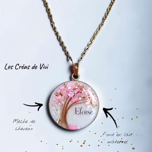 Può includere: Un ciondolo rotondo su una catenina dorata con sfondo bianco, un design ad albero realizzato con ciocche di capelli castani e accenti floreali rosa. Il nome Eloise è stampato in nero. Il testo include Les Créas de Vivi e Mèche de cheveux.