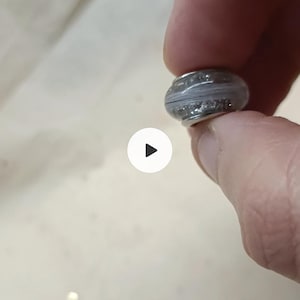 May include: A small, round bead with a silver-coloured metal base and a grey, textured centre. The bead has a band of sparkling silver accents. The bead is held between fingers.