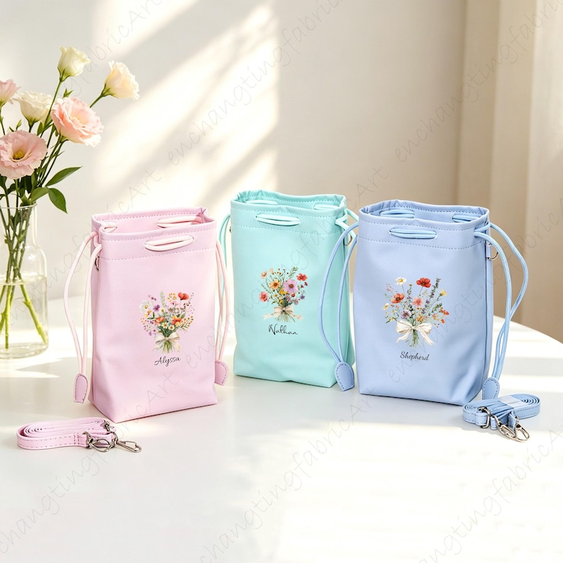 以下が含まれることがあります： パステルカラーの巾着バッグ3つ。各バッグには花のブーケのデザインと名前（Alyssa、Nathan、Shepherd）が施されています。バッグにはお揃いのストラップとシルバーカラーの金具が付いています。バッグは白い表面に置かれています。