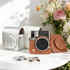 May include: A black and brown instant camera with a brown leather case, a silver case, and a white case. The silver case has the name "Roberta" engraved on it. Two instant photos are in front of the camera.