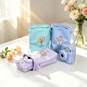 以下が含まれることがあります： パステルカラーのカメラバッグと水色のインスタントカメラのコレクション。バッグは青と紫の色合いで、それぞれに花のブーケのデザインと名前が飾られています。背景には花瓶に入った花があります。