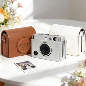 May include: A silver camera with a black lens is displayed with three leather cases. A brown case with a circular design and a white case are on either side of the camera. A small photo is in front of the camera.