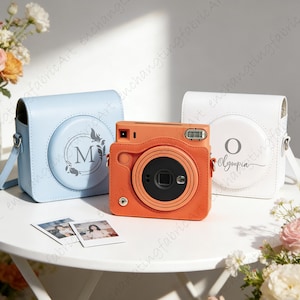 May include: An orange instant camera with a black lens is positioned between a light blue and a white camera case. The light blue case has the letter "M" and a floral design. The white case has the letter "O" and the name "Olympia". Two photos are in front.