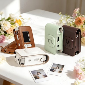 May include: Leather camera cases in brown, white, and mint green. The cases are designed for instant cameras, some with initials. Two instant photos are present, alongside floral arrangements. The cases are designed to fit instant cameras.