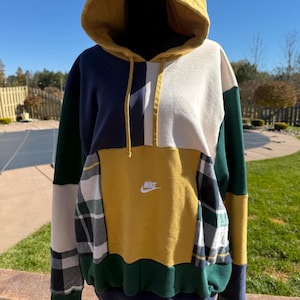 May include: A color-blocked hooded sweatshirt featuring a mustard yellow hood and central panel, navy blue, cream, and dark green sections, and plaid fabric accents. The Nike logo is embroidered in white on the center chest area.