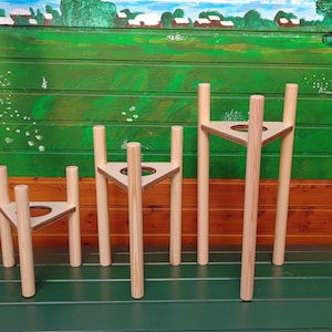 Op de afbeelding: Vier houten plantenstandaards in verschillende hoogtes, elk met een driehoekige basis en drie poten. De standaards zijn gemaakt van natuurlijk hout en hebben een lichtbruine kleur.